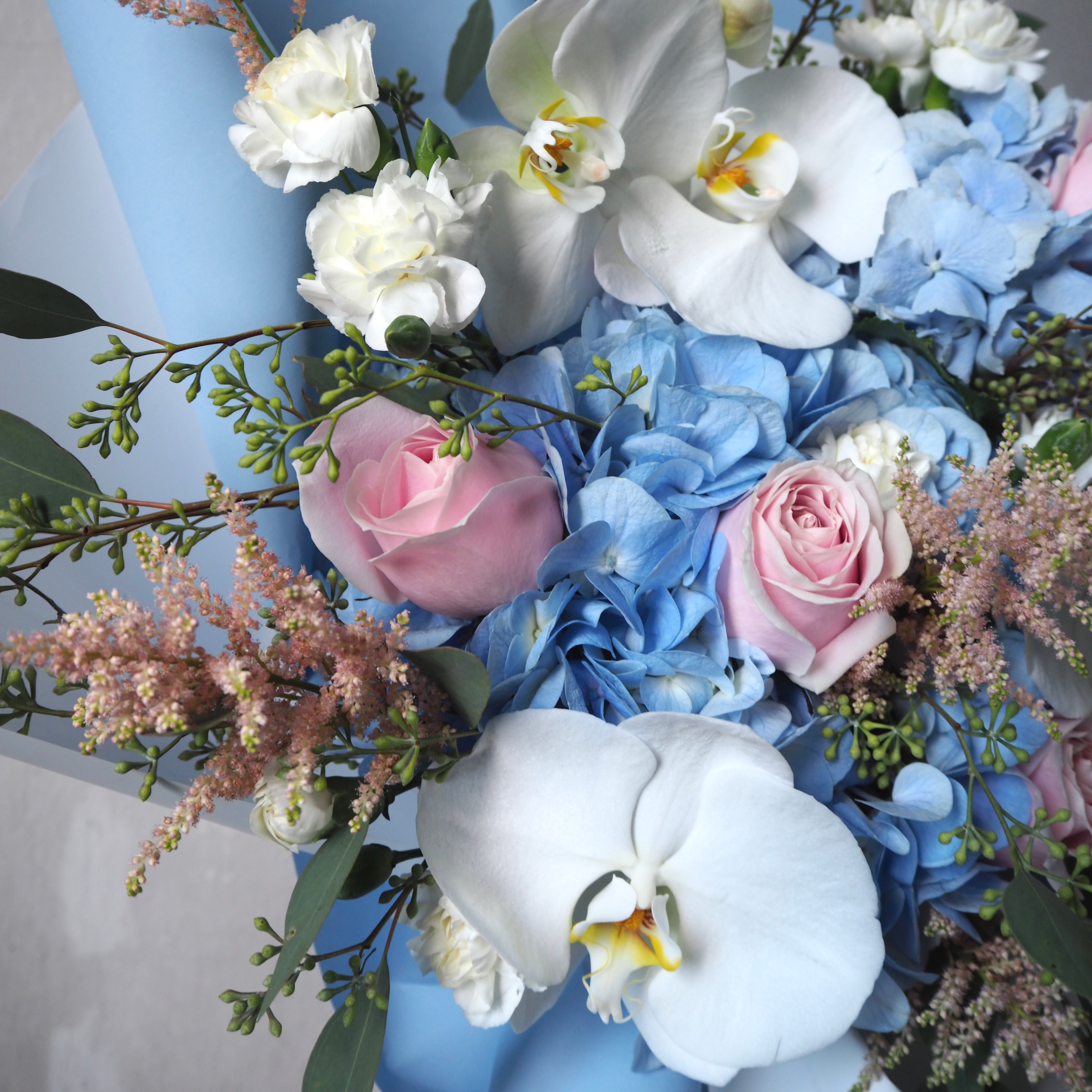 Bouquet of flowers including white orchids, pink roses, and blue hydrangeas on a light gray background.
