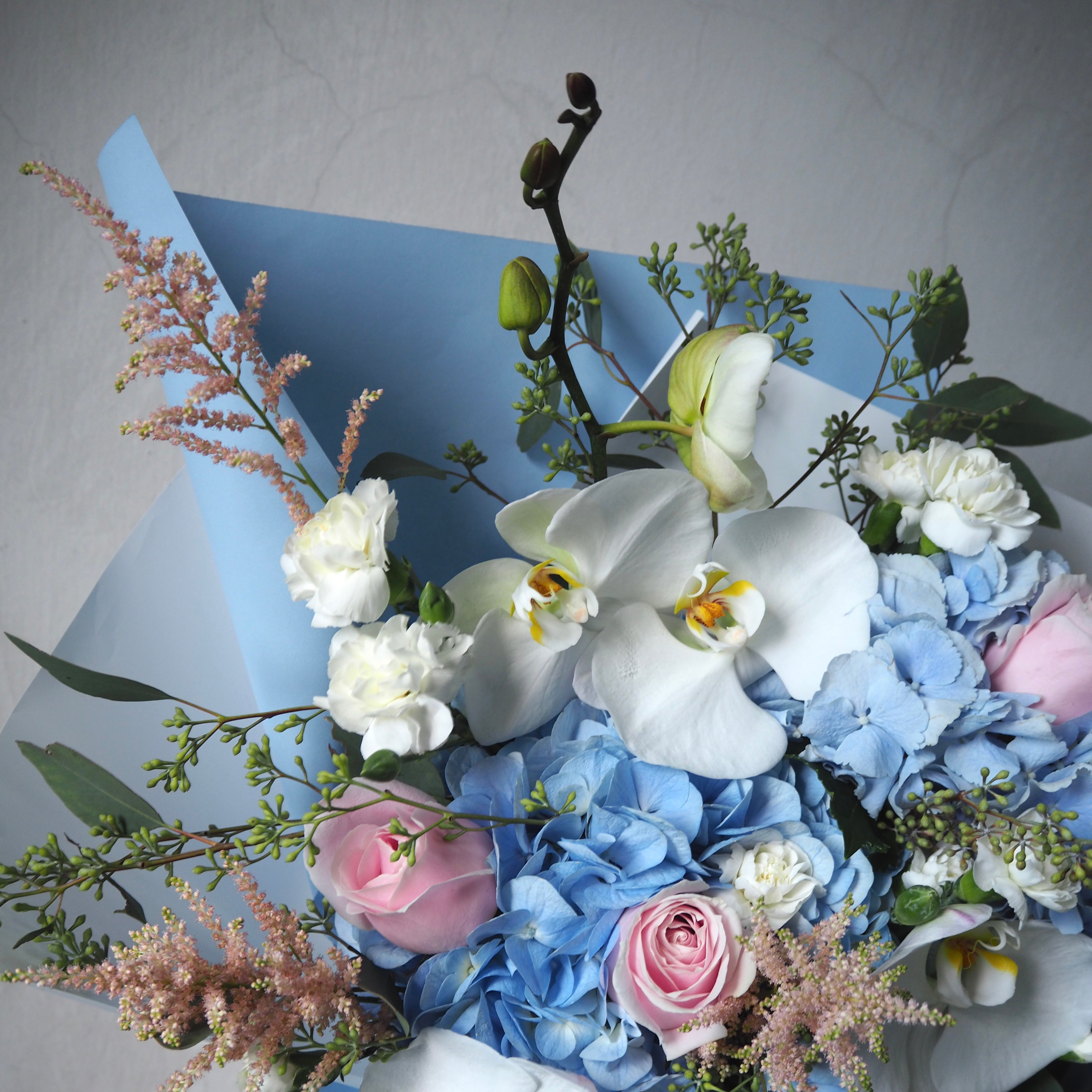Bouquet of flowers with white orchids, blue hydrangeas, and pink roses on a light gray background.