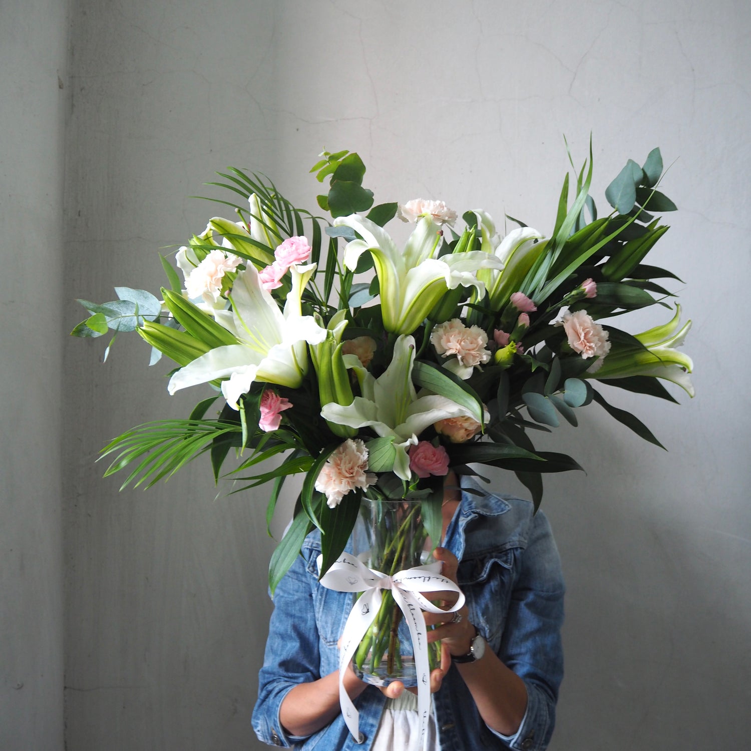Person holding a vase of white lily flowers against a plain background.