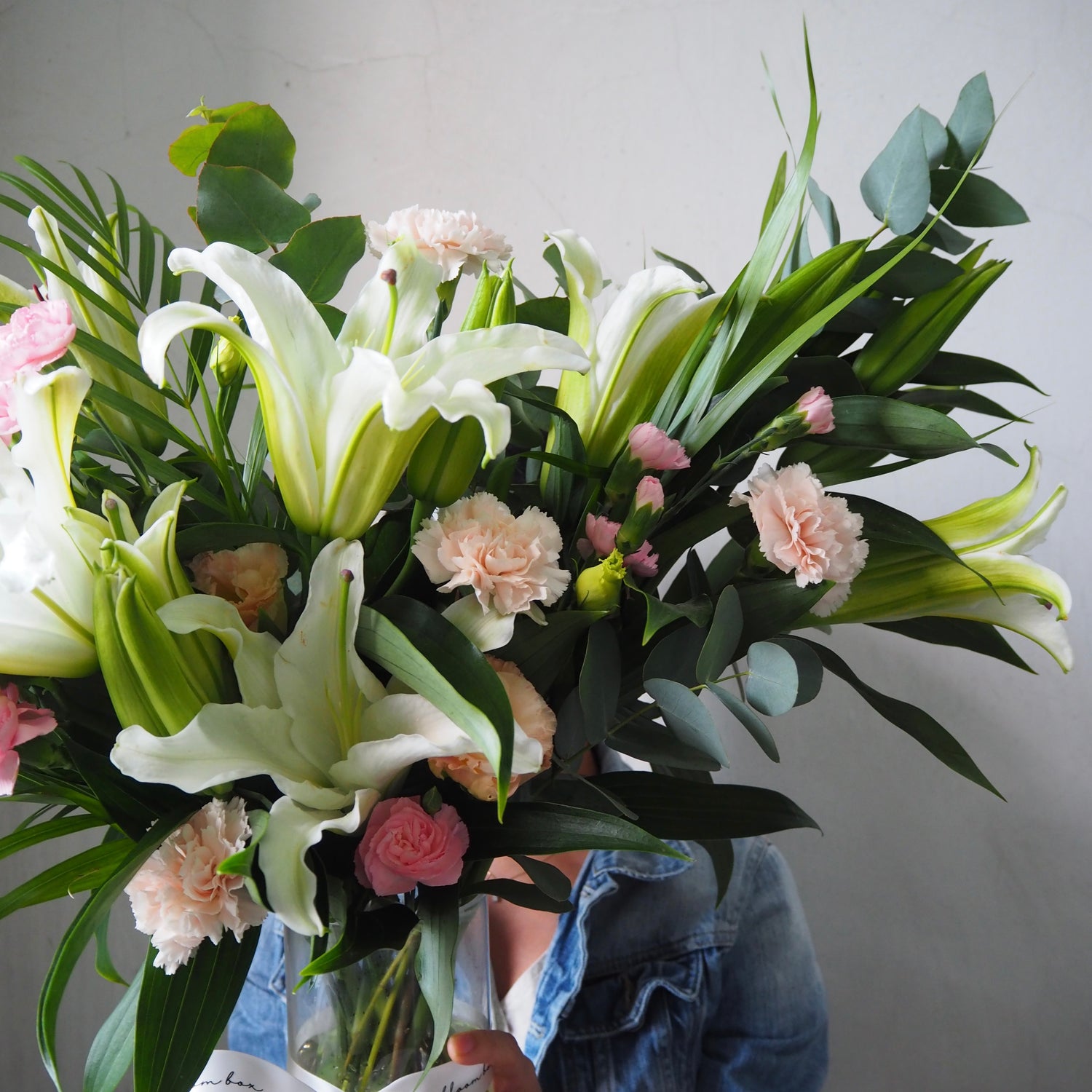 Bouquet of flowers with white lilies, pink carnations, and green leaves held by a person wearing a denim jacket.