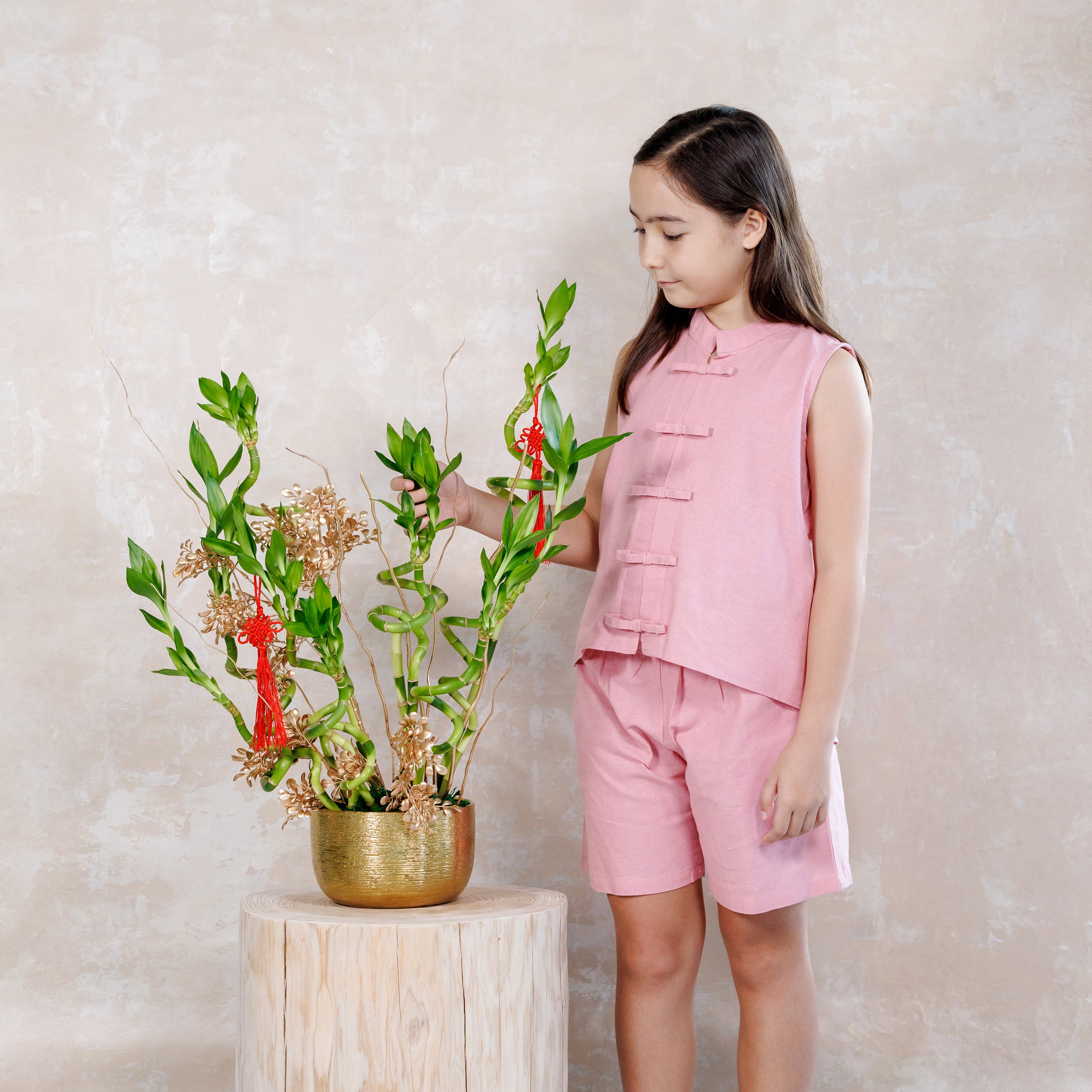 Young girl in a pink outfit standing next to a decorative plant on a small table.