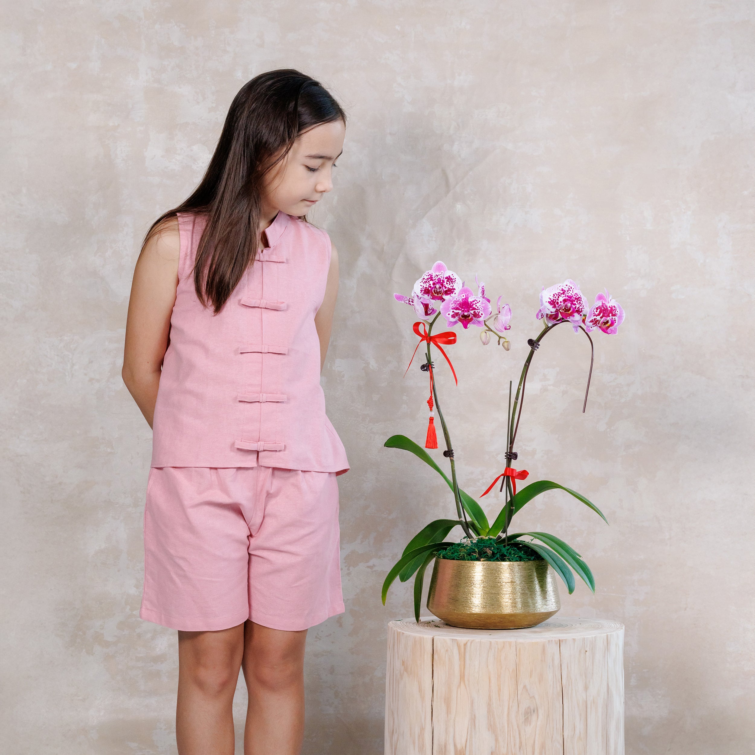 Young girl in pink outfit looking at a potted orchid plant on a wooden stool against a beige wall.