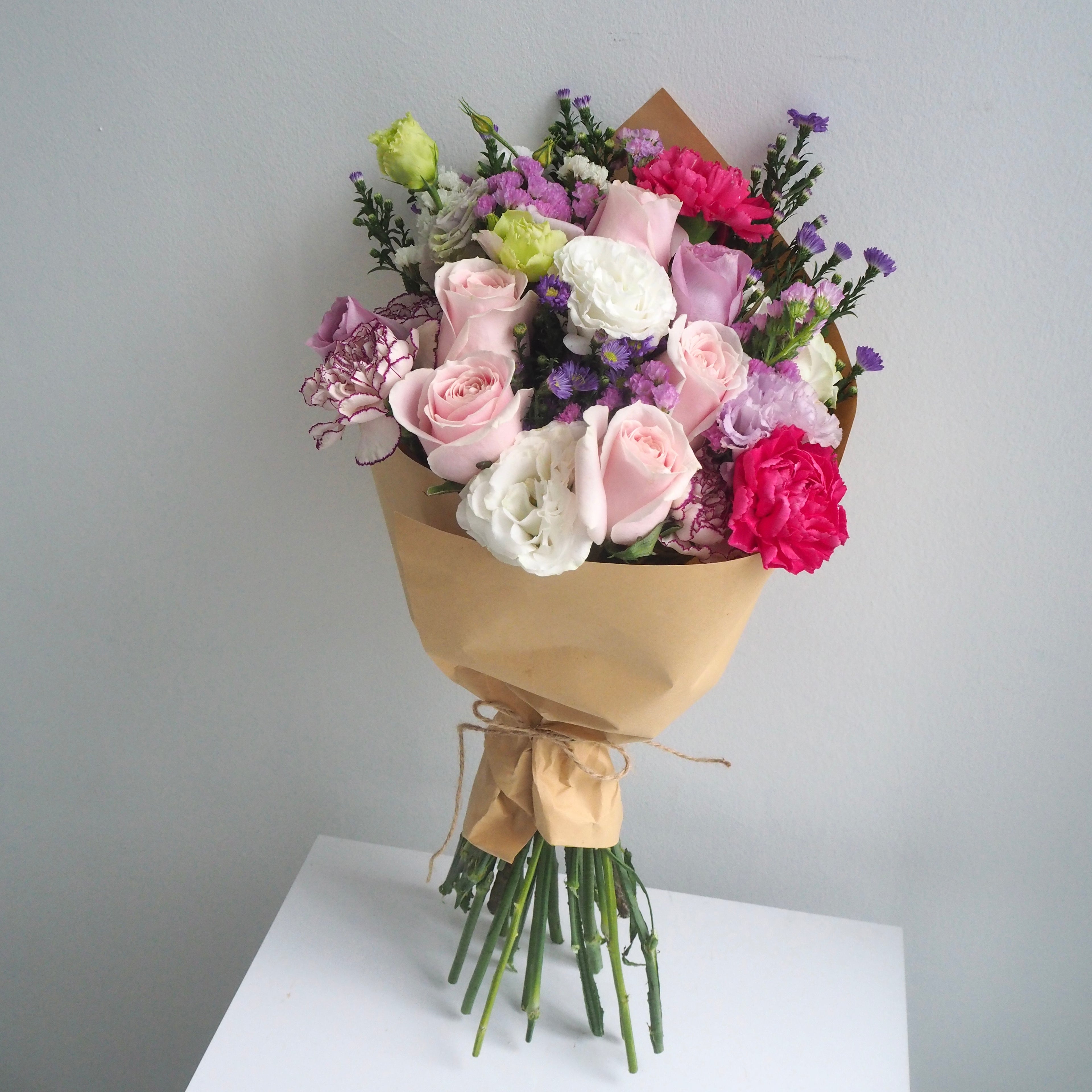 A bouquet of mixed flowers wrapped in brown kraft paper and tied with a ribbon, displayed on a white surface against a light gray background.