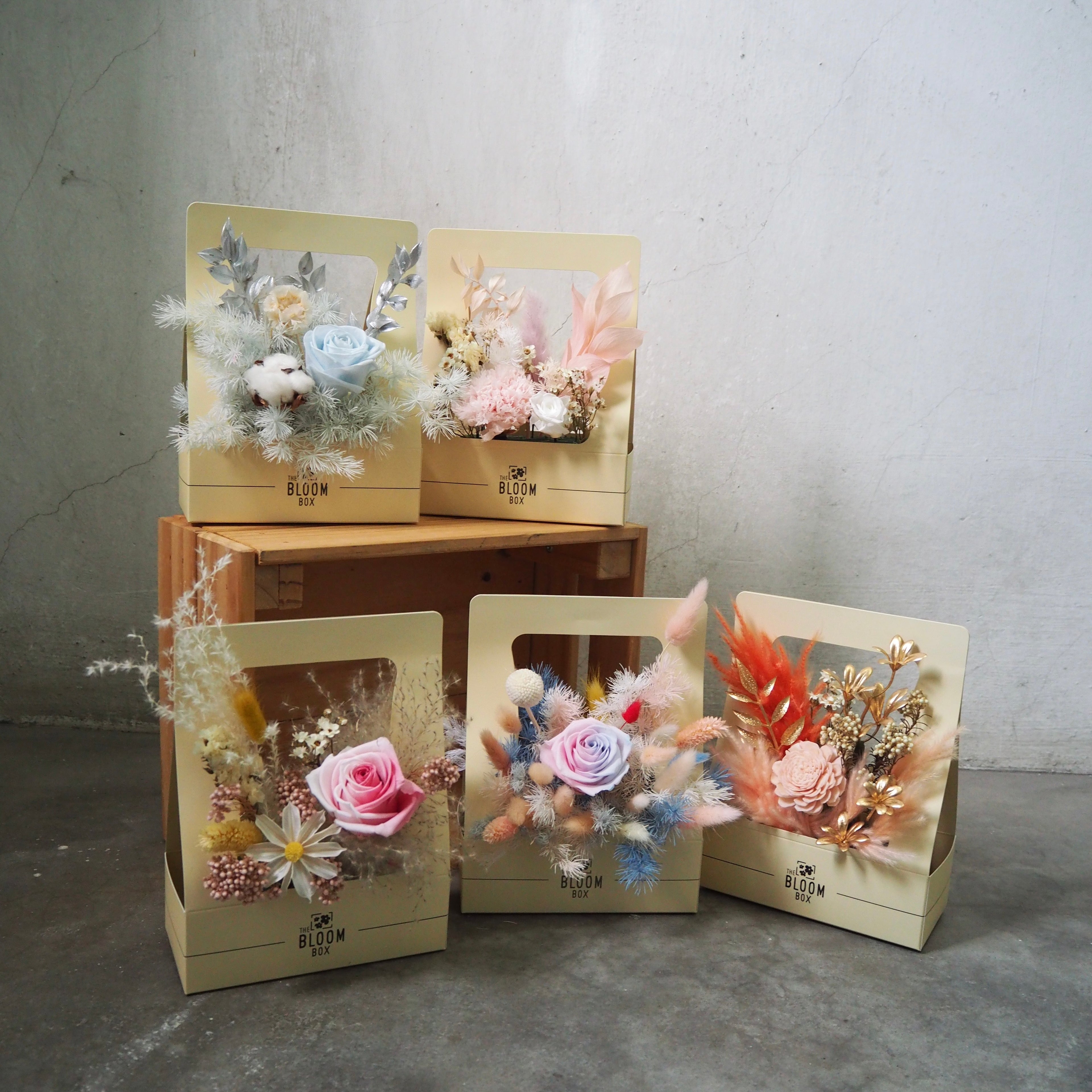 Decorative flower arrangements in transparent boxes on a concrete floor with a gray wall background.