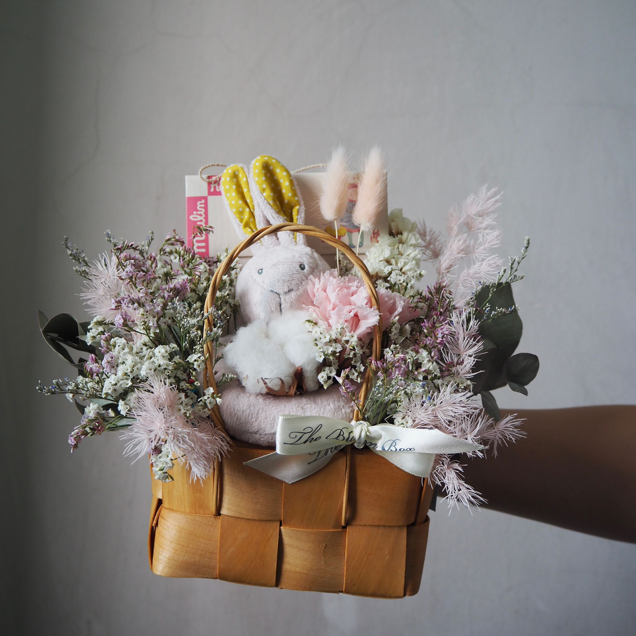 Braided basket with plush toy, flowers, and books against a gray background