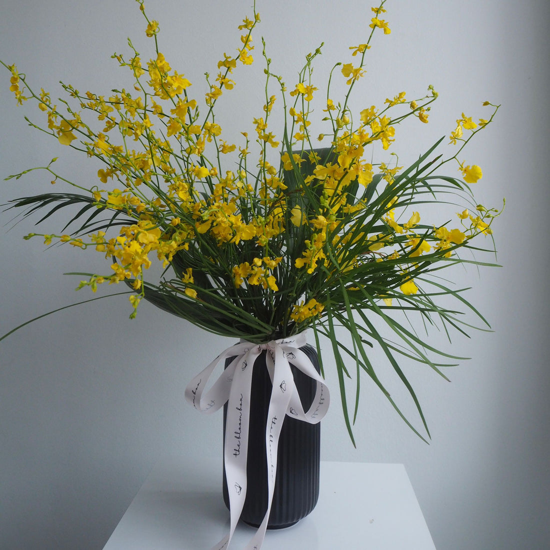 A large yellow orchid flower arrangement with green leaves and stems, tied with a white ribbon printed with the word &