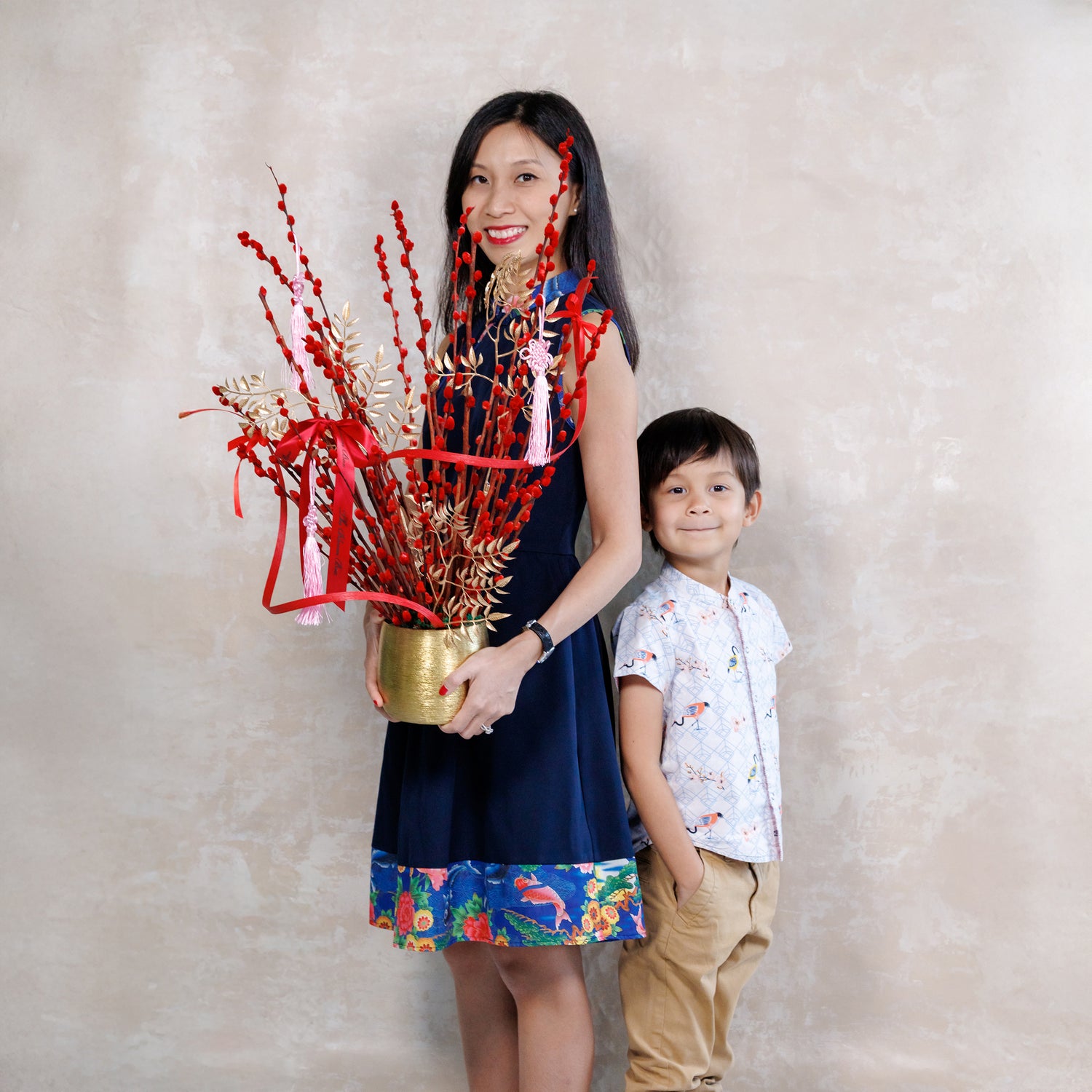 Woman holding a decorative plant with red ribbons and a child standing next to her against a plain background