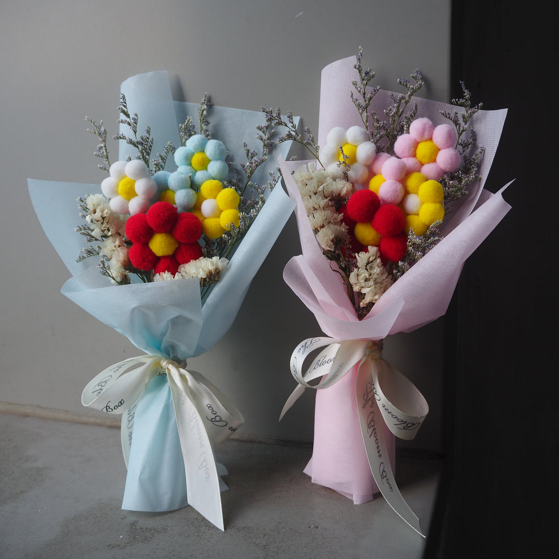 Two preserved flower bouquets with one featuring pink and white flowers and the other with blue and yellow flowers, both with a white ribbon.