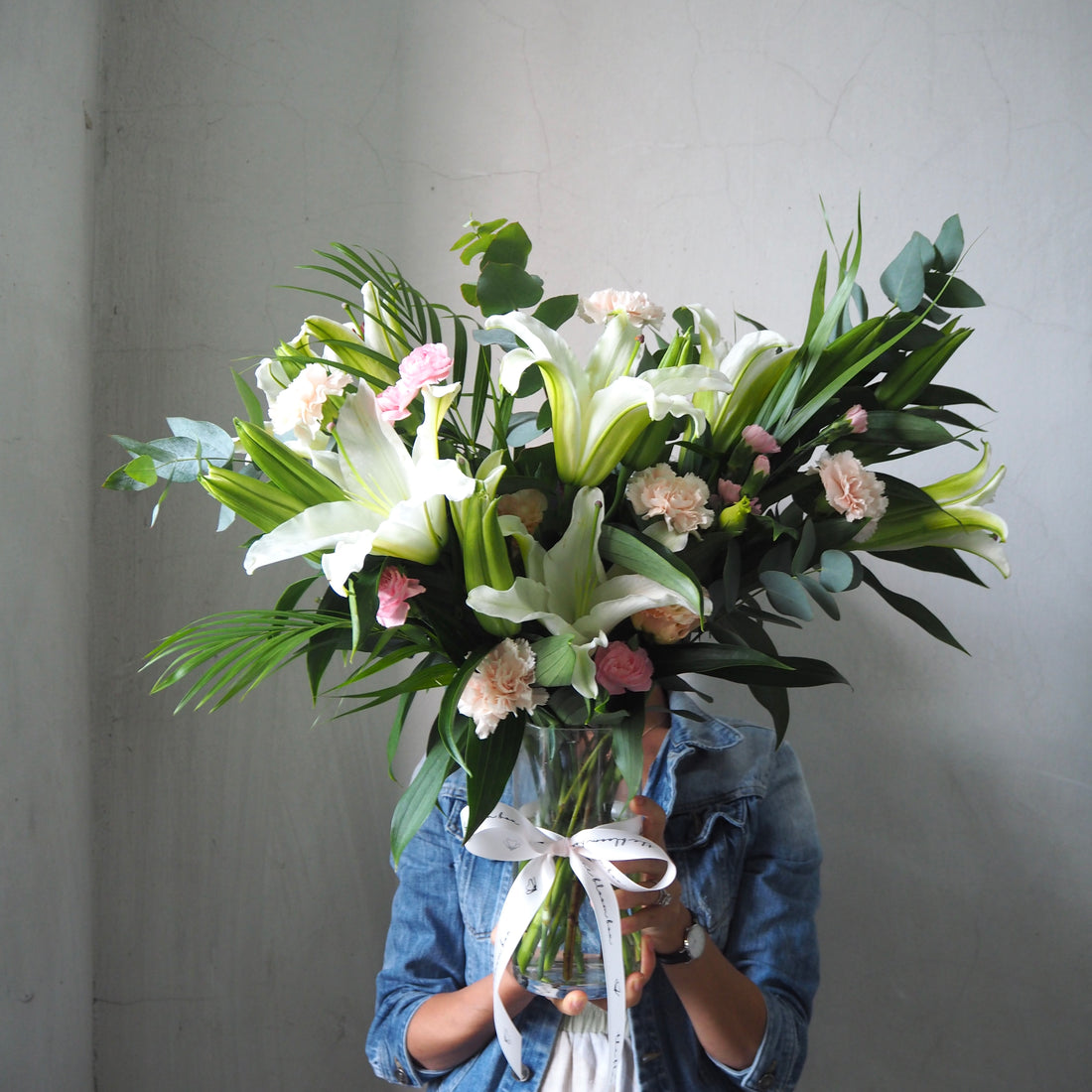 Person holding a vase of white lily flowers against a plain background.