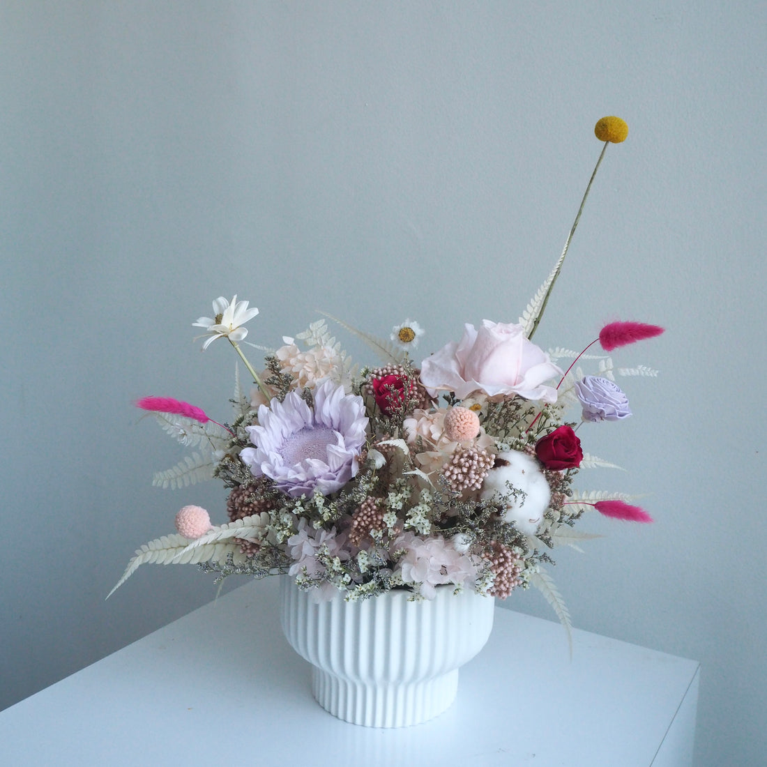 A dried flower arrangement in a white ribbed ceramic vase, placed on a white surface against a pale blue-gray wall. The bouquet includes soft pink, lavender, cream, burgundy, and yellow dried blooms with feathery accents.