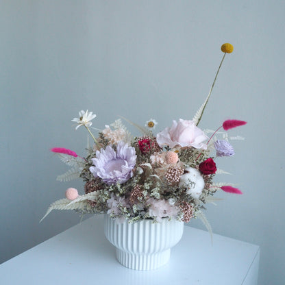 A dried flower arrangement in a white ribbed ceramic vase, placed on a white surface against a pale blue-gray wall. The bouquet includes soft pink, lavender, cream, burgundy, and yellow dried blooms with feathery accents.