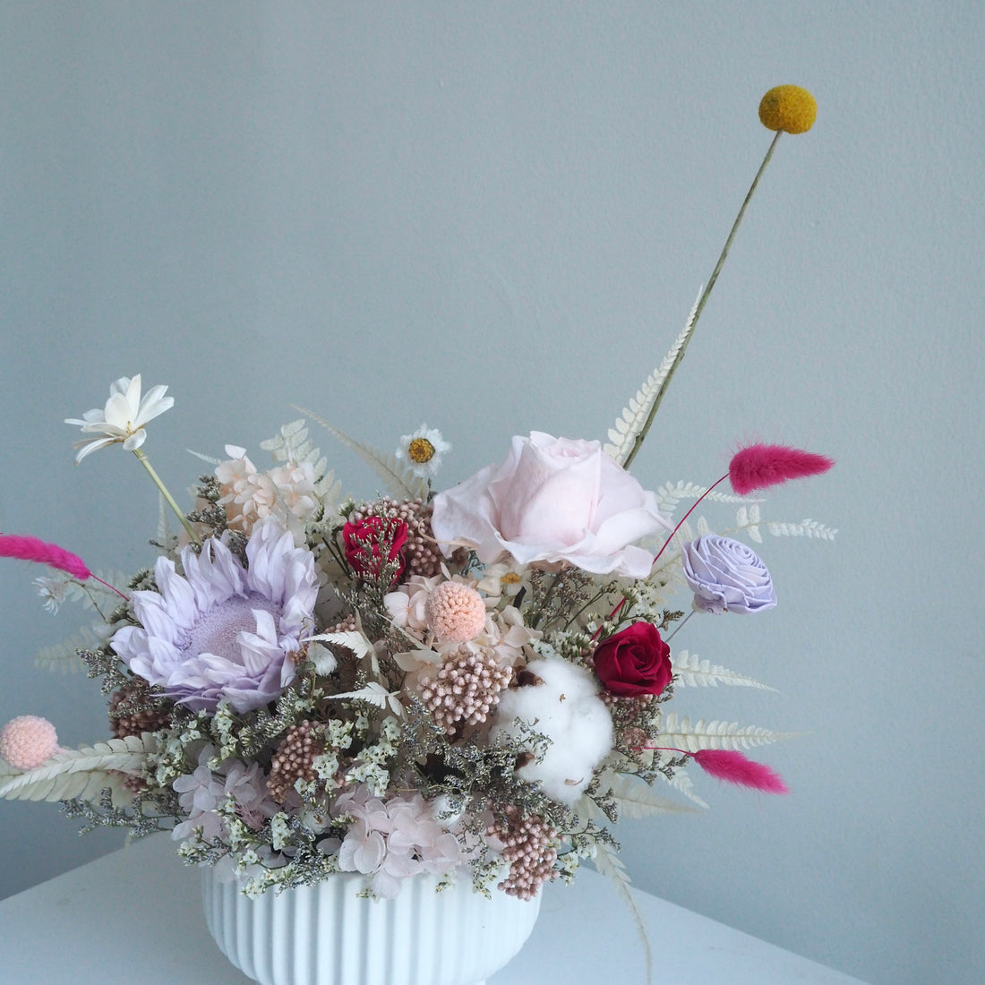 A dried flower arrangement in a white ribbed vase, featuring a large pale pink rose, a lavender protea, red roses, cotton boll, and various feathery and spherical dried flowers, set against a plain light blue-gray wall.
