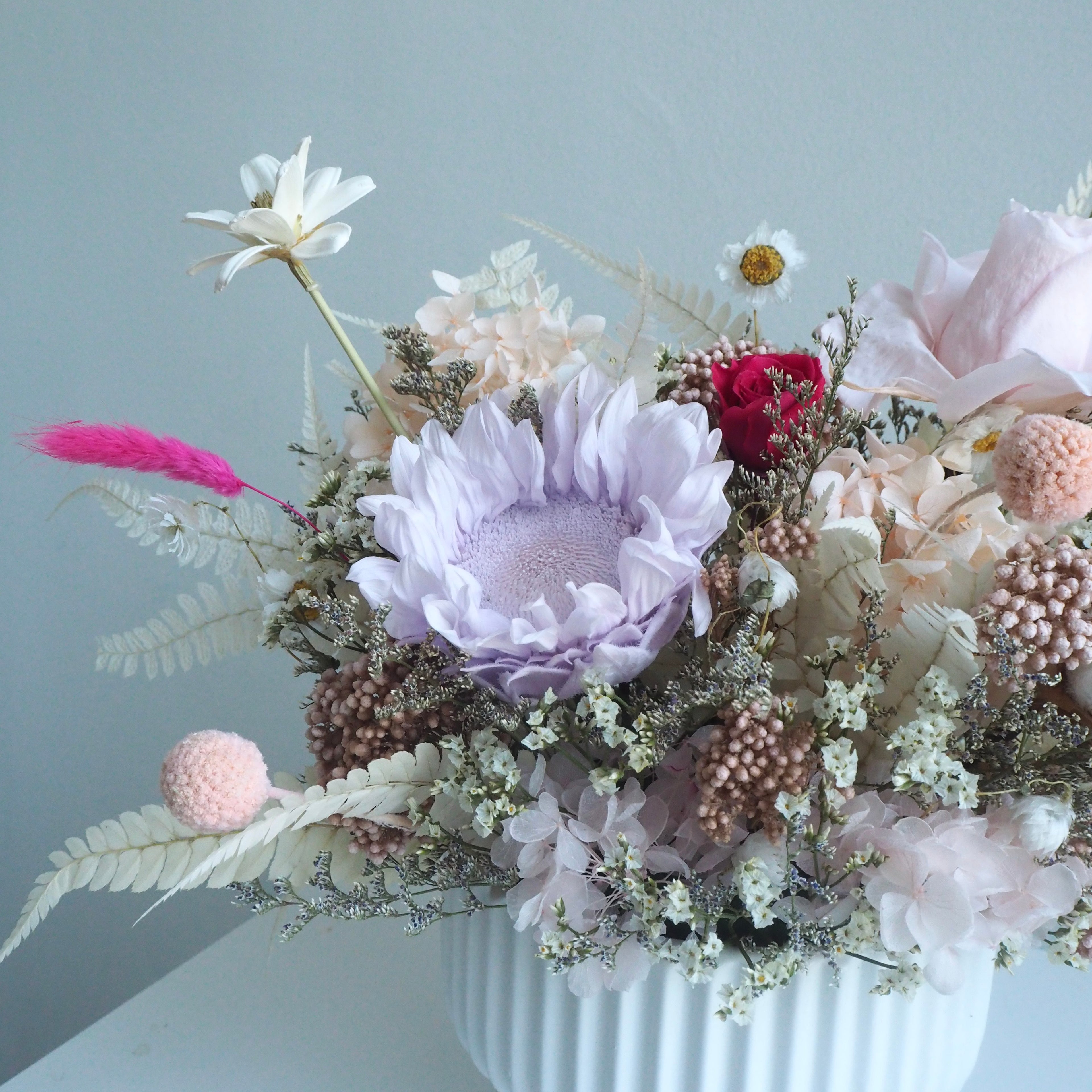 A dried flower arrangement in a white ribbed vase, featuring a large light purple protea, pale pink flowers, white daisies, and soft beige foliage against a pale blue-gray background.