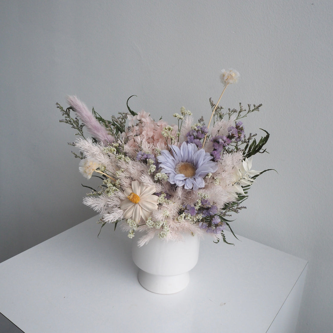 A dried flower arrangement in a white ceramic pot, featuring pale pink, lavender, and white blossoms with green foliage, set on a white surface against a light gray wall.