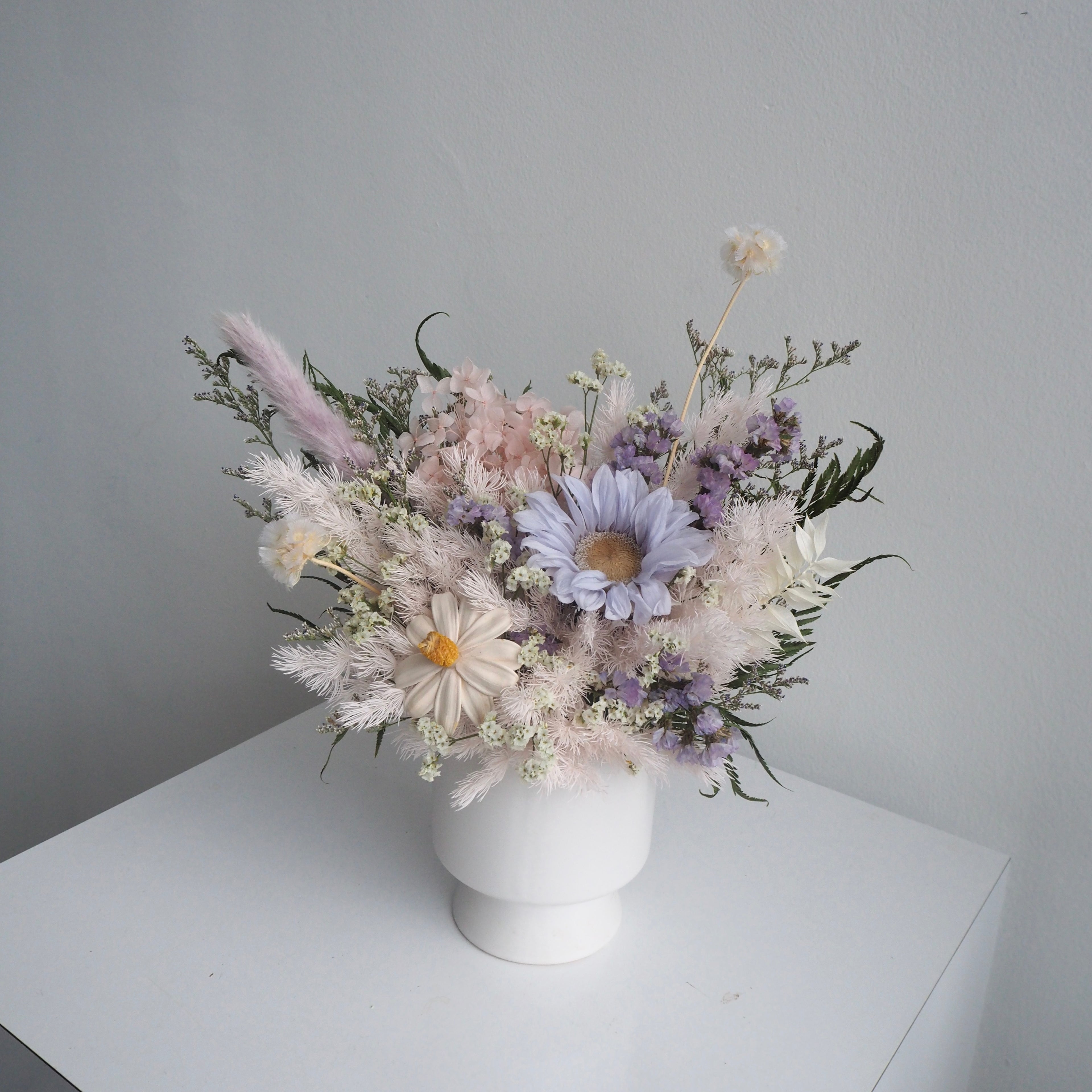 A dried flower arrangement in a white ceramic pot, featuring pale pink, lavender, and white blossoms with green foliage, set on a white surface against a light gray wall.