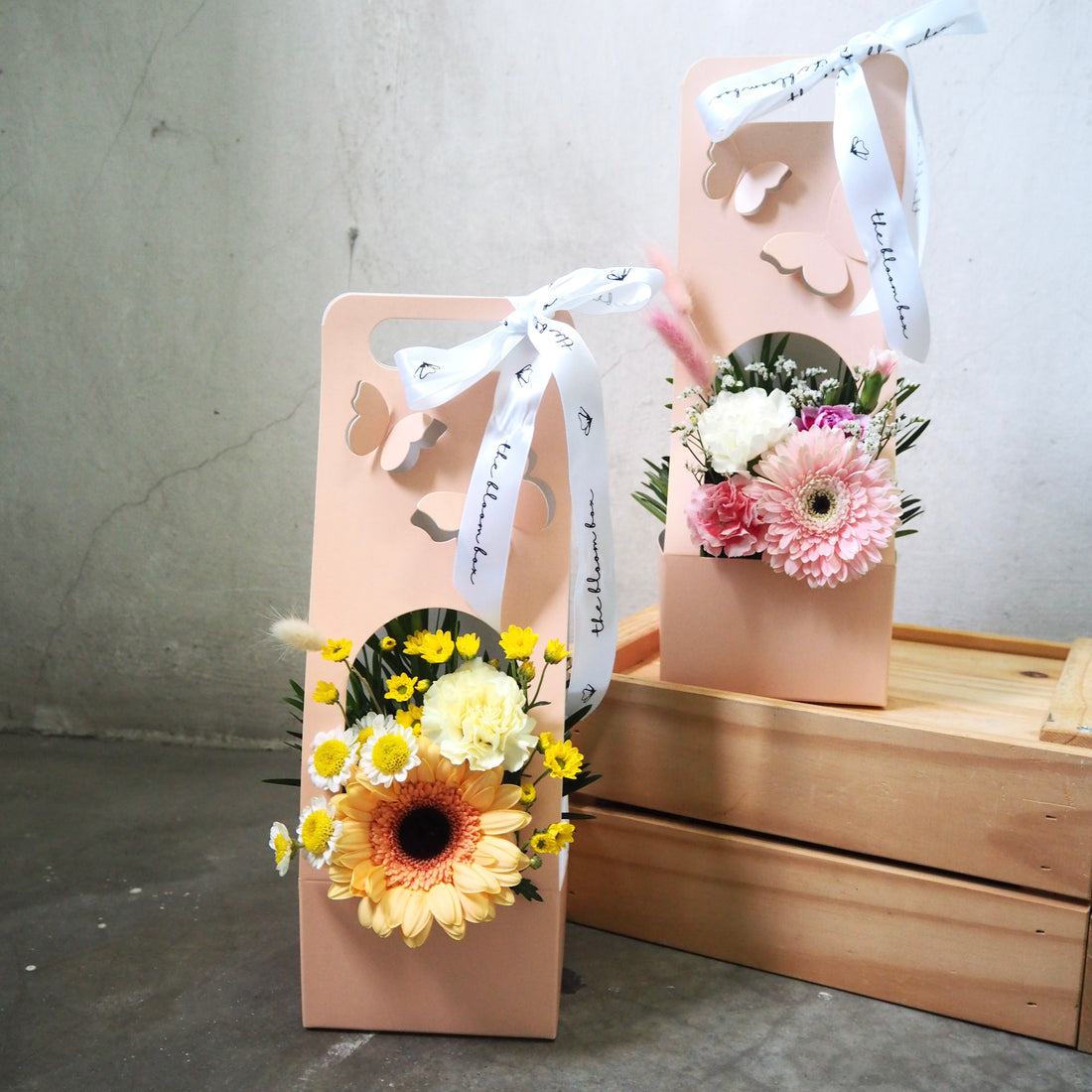 Two floral arrangements in decorative boxes with ribbons on a wooden surface.