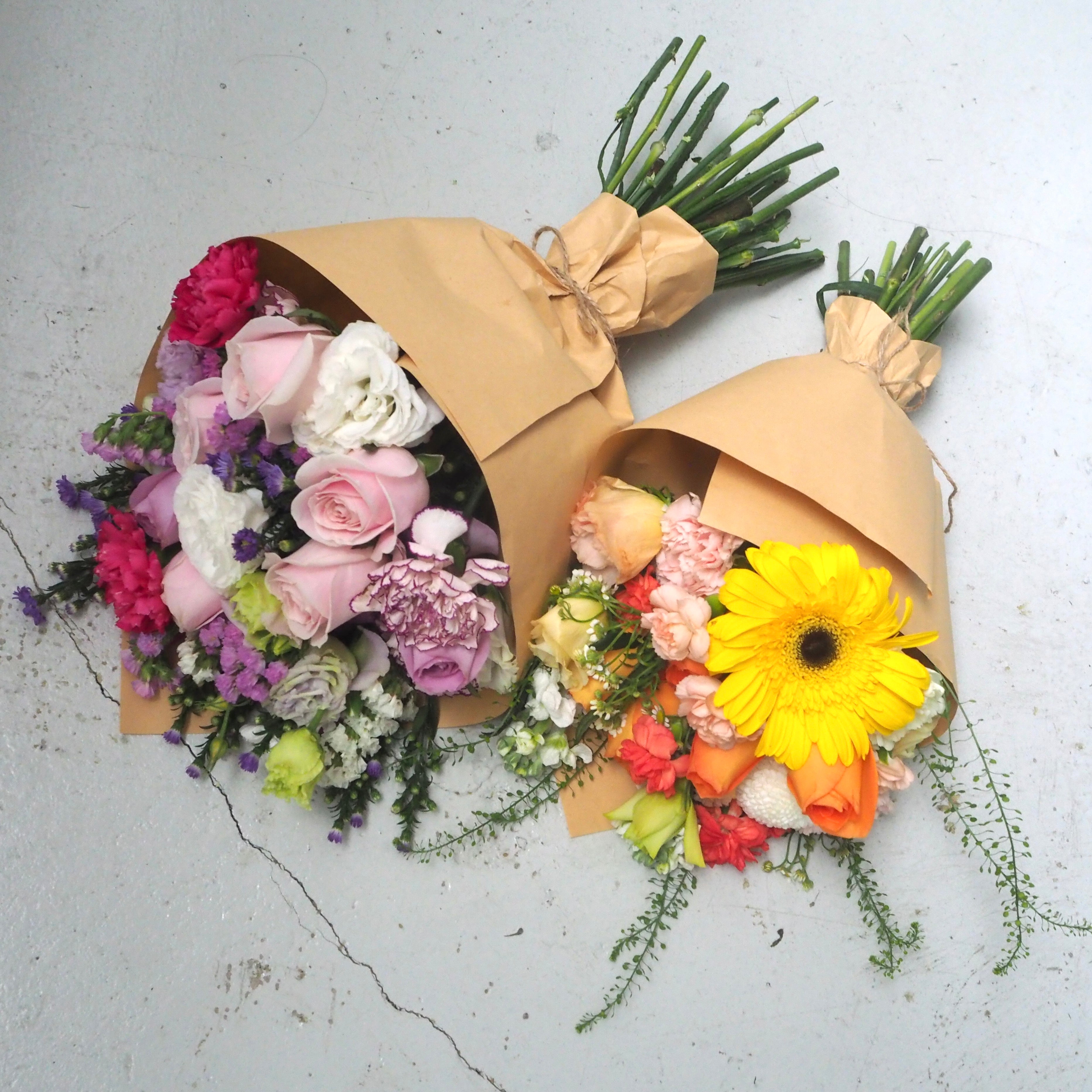 Two wrapped flower bouquets with green stems, wrapped in brown paper, displayed on a light gray concrete surface. One bouquet features pink and purple roses, lavender, and white flowers.