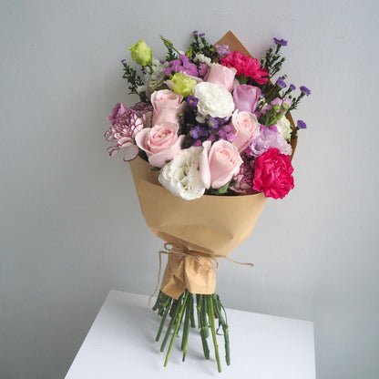 A bouquet of mixed flowers wrapped in brown kraft paper and tied with a ribbon, displayed on a white surface against a light gray background.