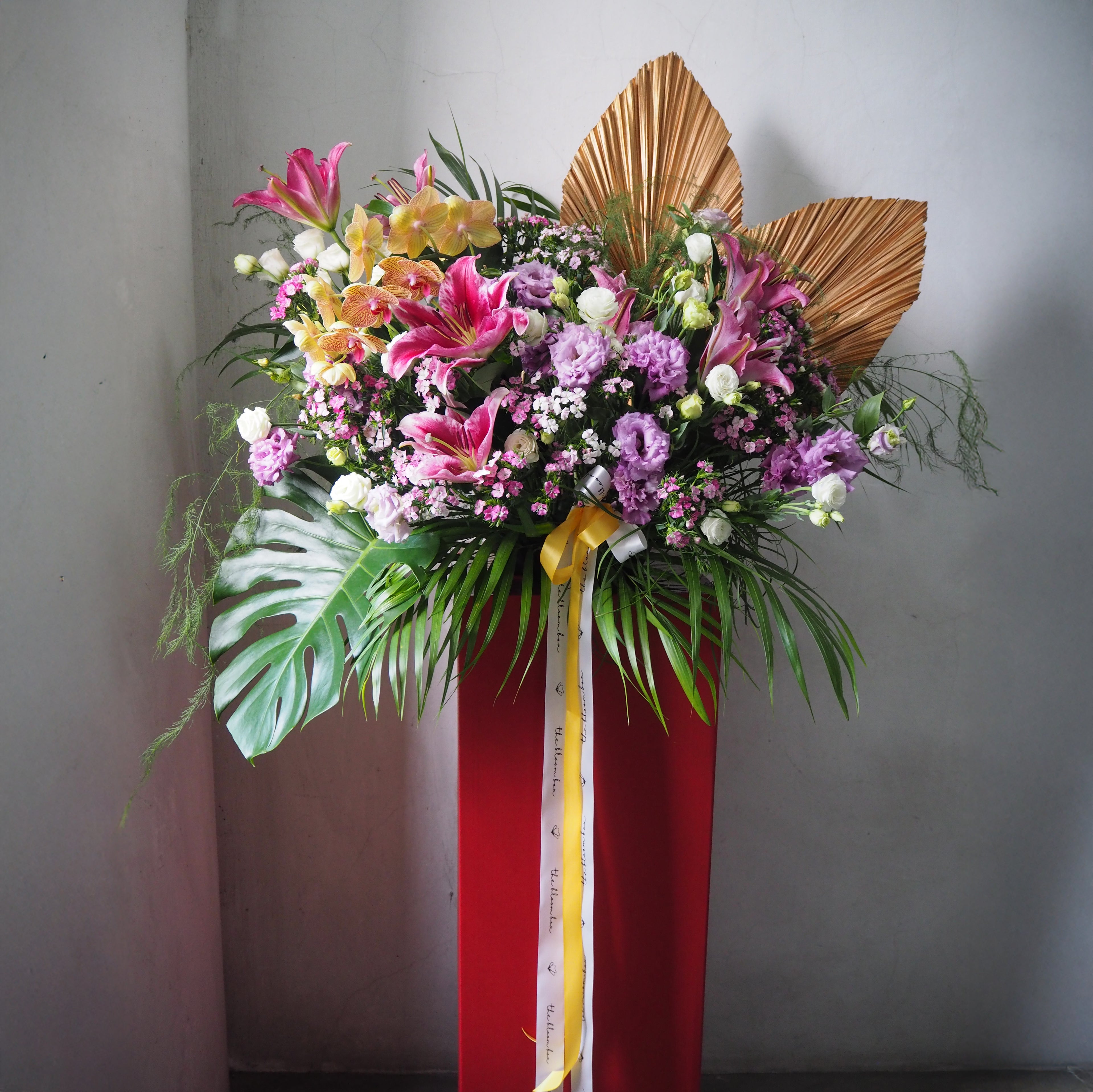 Colorful floral arrangement on a red stand with a gold palm leaf decorative element against a gray background