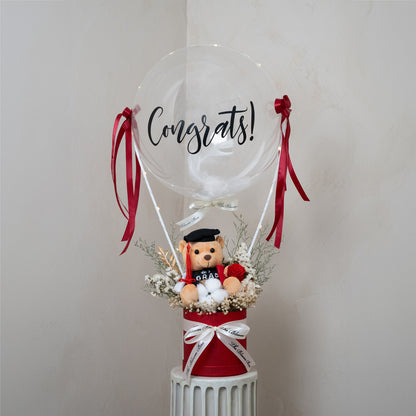 grad bear in red box with red preserved flower arrangements and congrats message balloon decorated with fairy light