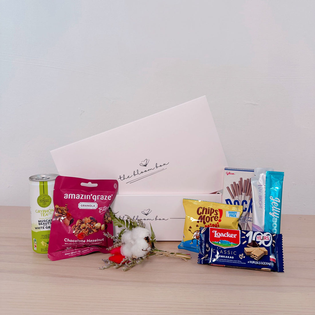 Assorted snacks and a small bouquet of flowers in front of a branded box on a wooden surface.