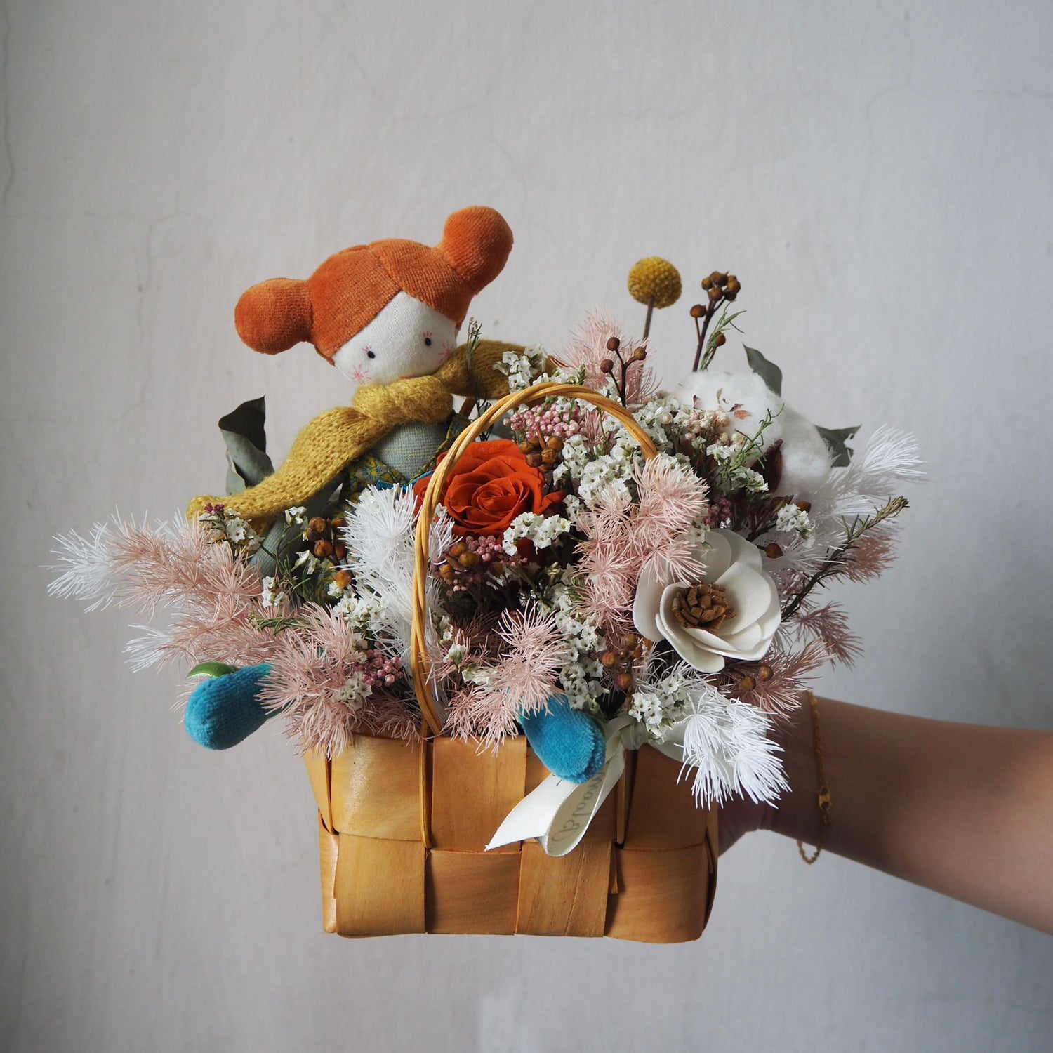 Floral arrangement in a woven basket held by a person, with a small plush toy wearing a red hat and yellow scarf.