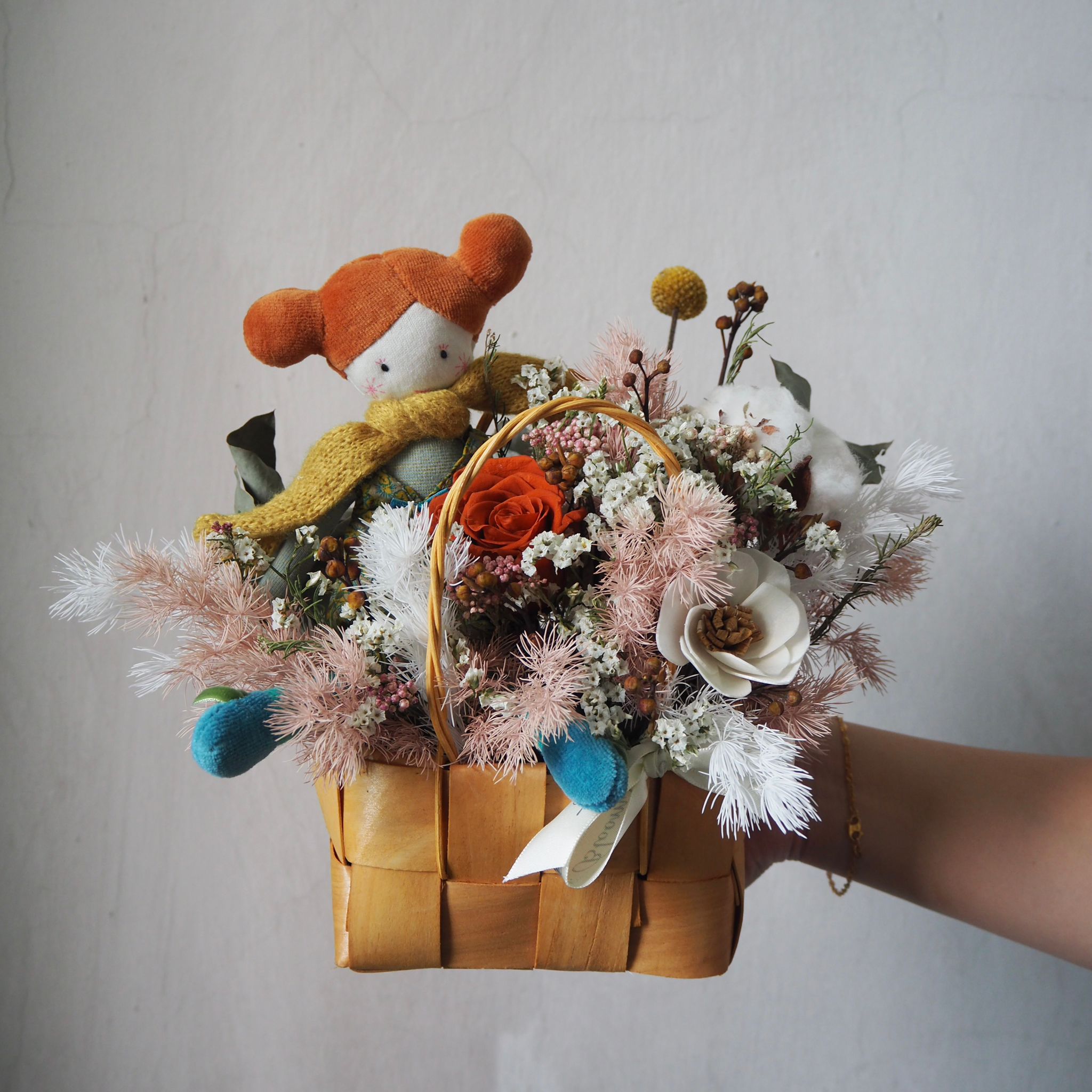 Floral arrangement in a woven basket held by a person, with a small plush toy wearing a red hat and yellow scarf.