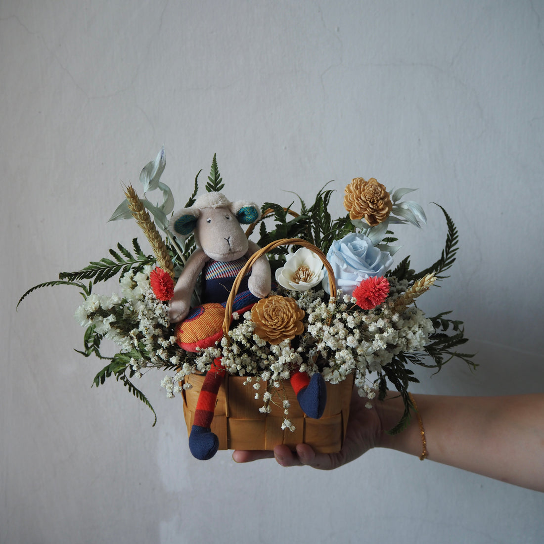 Hand holding a decorative arrangement with flowers, a small plush toy, and a basket against a plain background.
