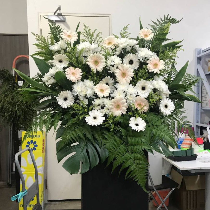 A floral stand with a serene blend of white chrysanthemums, gerberas, and green foliage, arranged to express sympathy for loss.