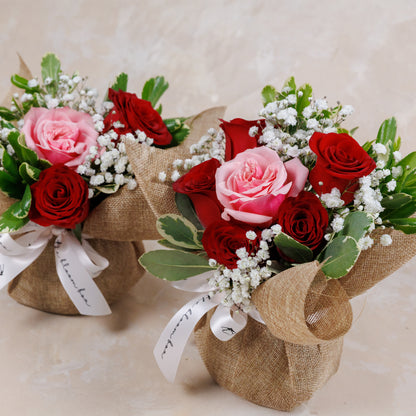 Two small bouquets of red and pink roses with greenery, wrapped in burlap and tied with white ribbons on a light background.