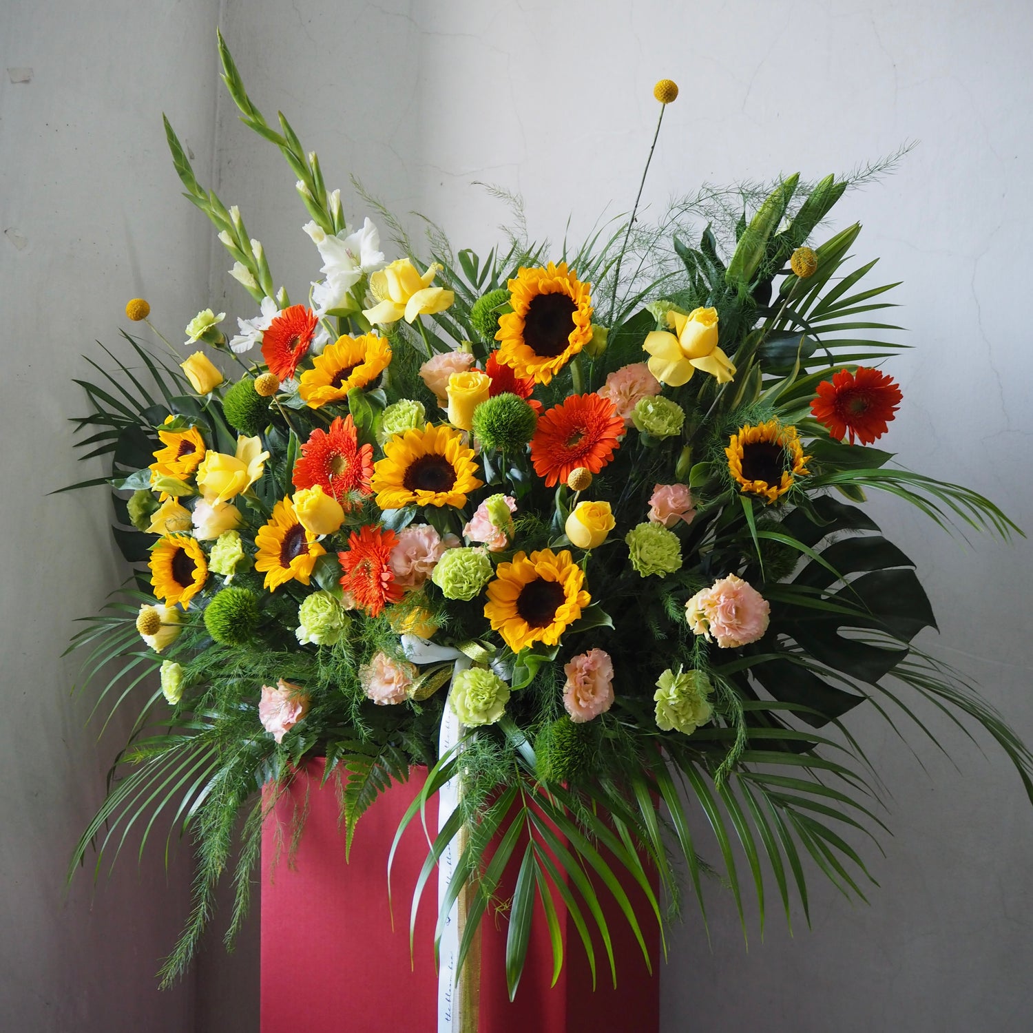 Colorful flower arrangement with sunflowers, daisies, and other flowers in a red container against a light gray background.
