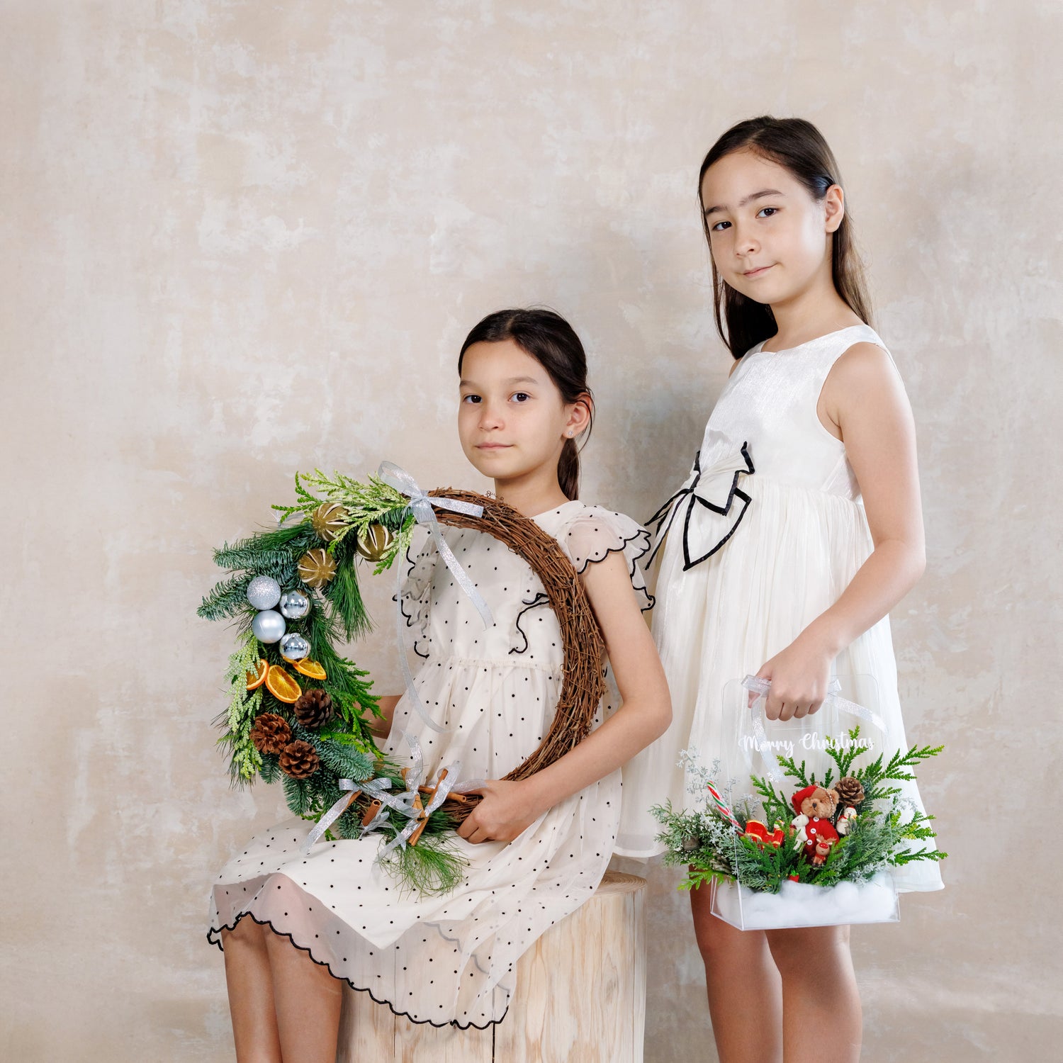 Two young girls in white dresses holding decorative wreaths against a plain background